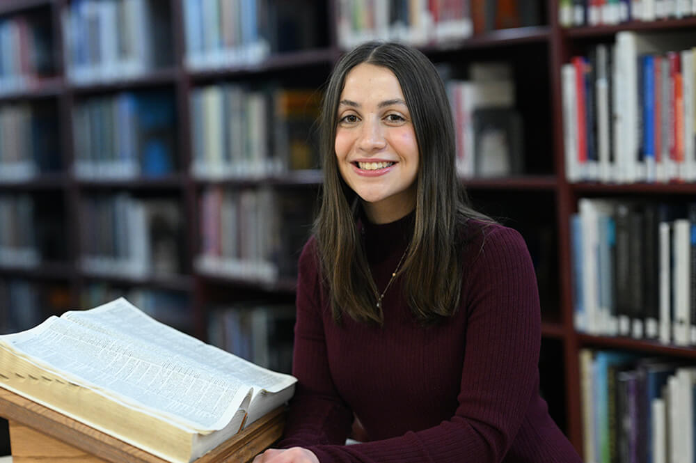 Ella Morris, female student with long brown hair in library with open book next to her and bookcases behind her