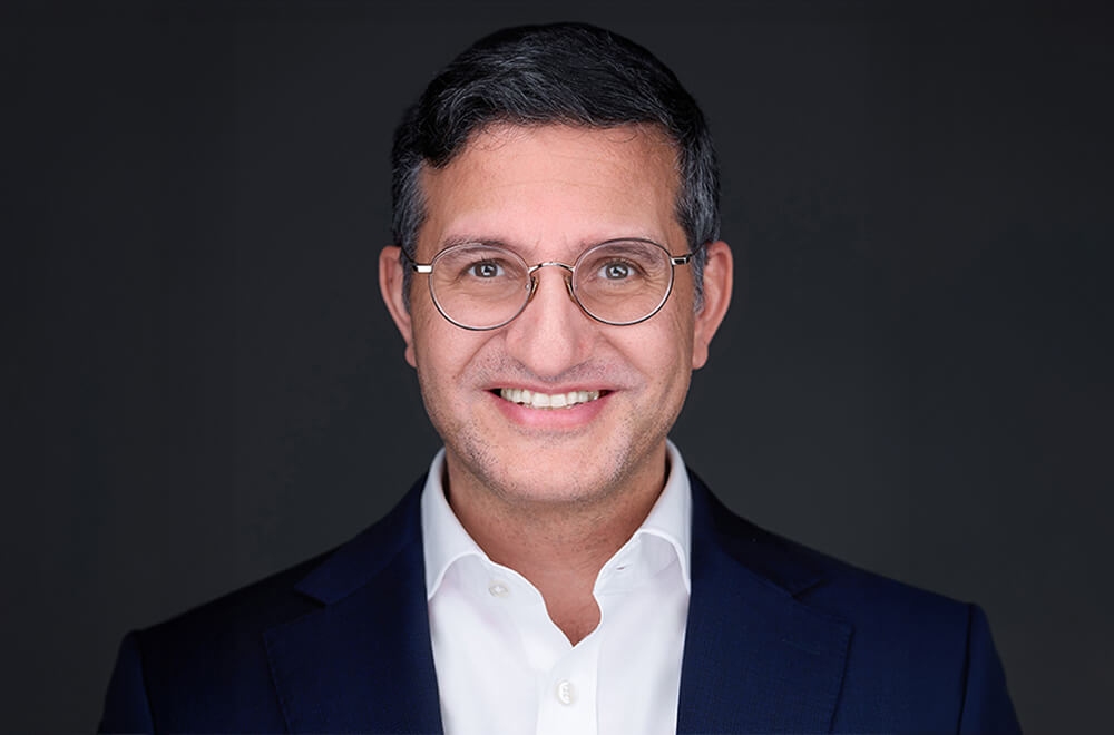 headshot of Saul Bakst, a smiling man with round rimmed glasses, a white shirt and dark suit jacket